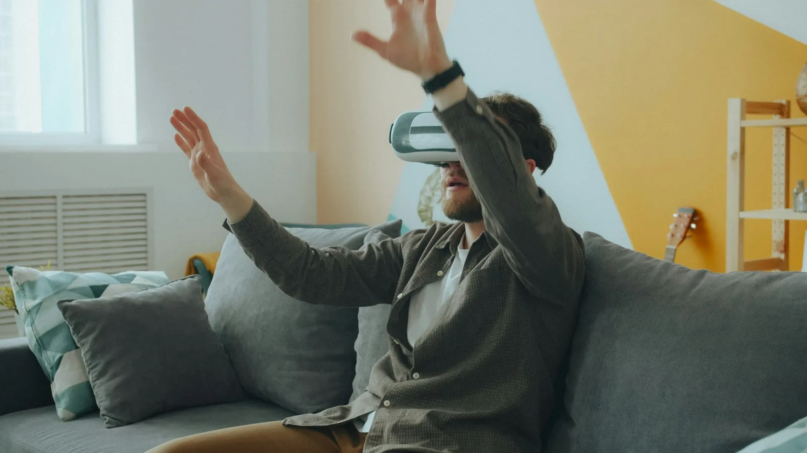 A man on a grey sofa wearing a headset, hands raised as if interacting with a virtual screen